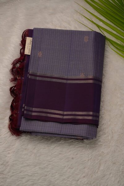 Folded light lavender lightweight Pattu saree showcasing fine zari checks, small floral buttis, and a contrast deep magenta gap border framed by metallic stripes.