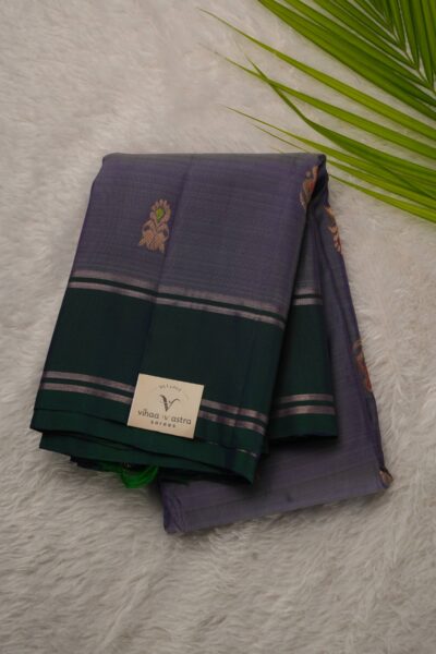 Folded lavender lightweight Pattu saree showcasing gold zari Meenakari buttis and a contrast bottle green gap border framed by metallic stripes.