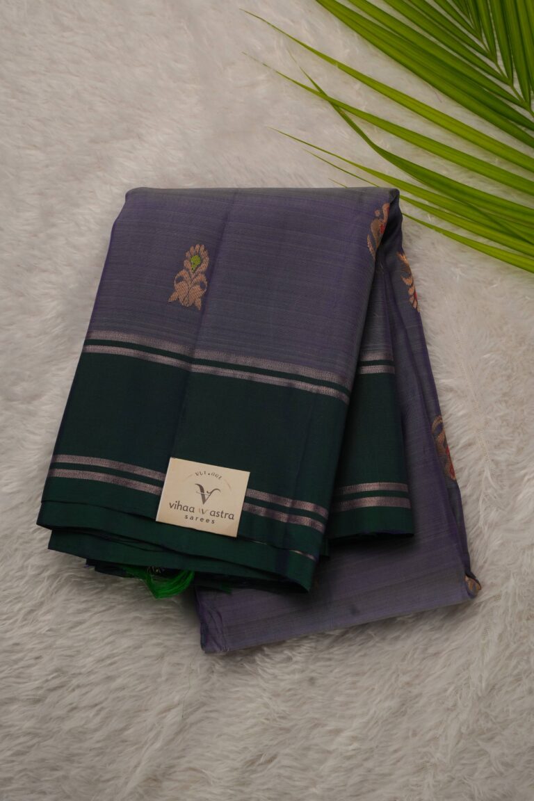 Folded lavender lightweight Pattu saree showcasing gold zari Meenakari buttis and a contrast bottle green gap border framed by metallic stripes.