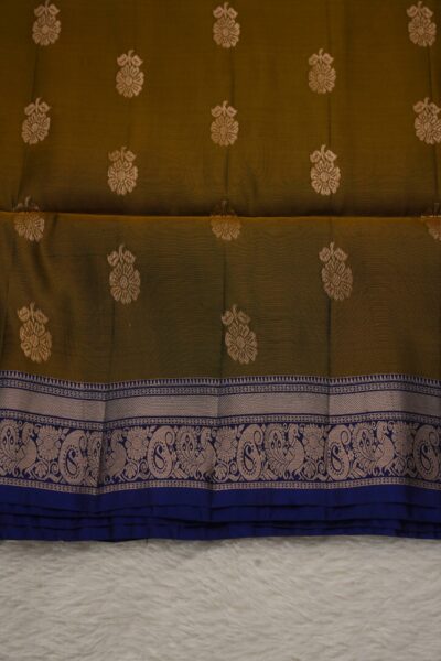 Close-up texture of an earthy mustard lightweight Pattu saree with large floral metallic buttis, alongside a beautifully detailed deep blue gold zari border featuring peacocks and paisleys.