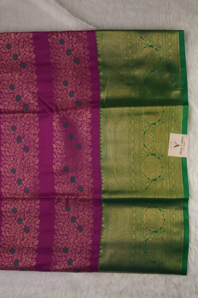 Close-up texture of the magenta blended silk body showing vertical gold zari lines alongside the heavy, intricate gold zari peacock work of the green Kanchi border.