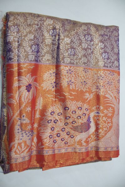 Folded purple Semi Kanchi Pattu saree with floral zari brocade, paired with a vibrant orange pallu displaying a beautifully woven scenic landscape featuring a grand peacock, deer, and trees with blue Meenakari accents.