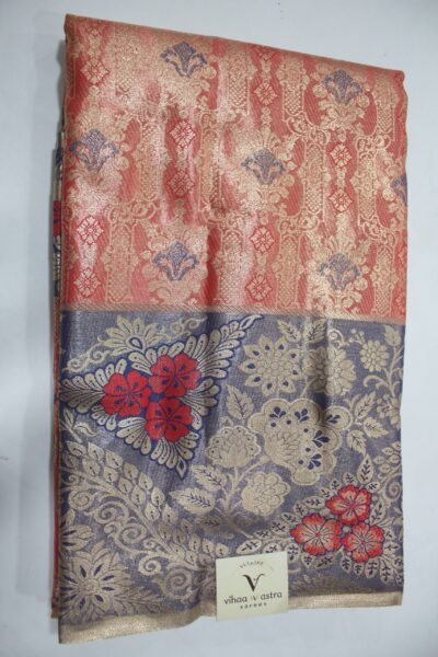 Folded red Semi Kanchi Pattu saree showcasing an all-over gold zari floral brocade with blue accents, and a heavy contrasting navy blue border featuring large floral motifs accented with bright red Meenakari weaving.