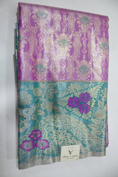 Folded violet Semi Kanchi Pattu saree showcasing an all-over gold zari floral brocade with green accents, and a heavy contrasting teal border featuring large floral motifs accented with bright violet Meenakari weaving.