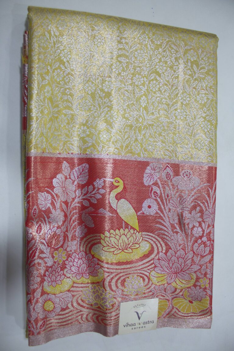 Folded yellow Semi Kanchi Pattu saree showing an all-over zari botanical brocade and a heavy contrasting coral-red border featuring a woven landscape of a swan in a lotus pond accented with yellow Meenakari weaving.