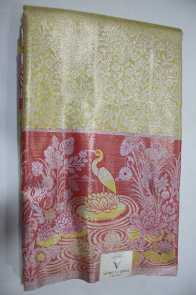 Folded yellow Semi Kanchi Pattu saree showing an all-over zari botanical brocade and a heavy contrasting coral-red border featuring a woven landscape of a swan in a lotus pond accented with yellow Meenakari weaving.