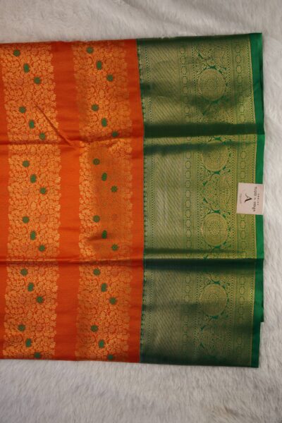 Close-up texture of the orange blended silk body showing vertical gold zari lines alongside the heavy, intricate gold zari peacock work of the green Kanchi border.