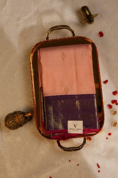 Top view of the beautifully folded peach pure handloom Kanjeevaram Pattu saree resting on a decorative brass tray, showcasing the delicate gold buttis and striking magenta gap border.