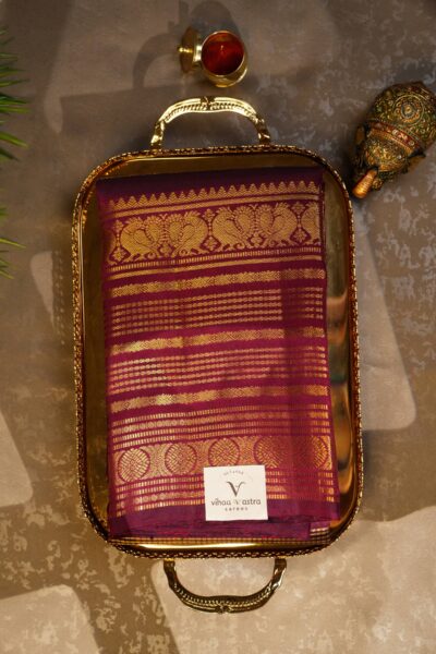 Beautifully folded self maroon pure handloom Kanjeevaram Pattu saree resting on a decorative tray, showcasing the delicate gold stripes and heavy border.