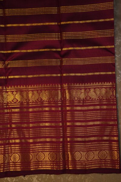 Close-up texture of the pure handloom maroon silk body showing the woven vintage gold zari lines alongside the heavy, intricate gold work of the Kanchi border.