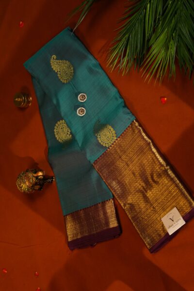 Top view of the beautifully folded tea green pure handloom Kanjeevaram Pattu saree resting on a decorative brass tray, showcasing the delicate gold buttis and heavy maroon border.