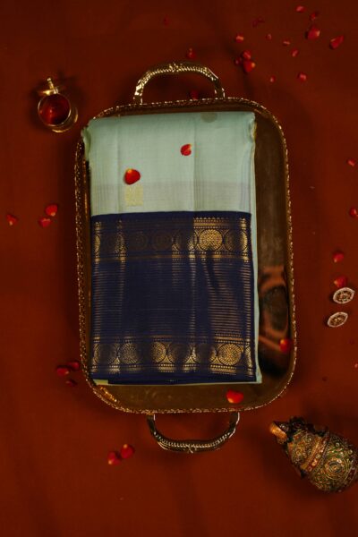 Top view of the beautifully folded light pista green pure handloom Kanjeevaram Pattu saree resting on a decorative tray, showcasing the delicate gold stripes and heavy navy blue border.
