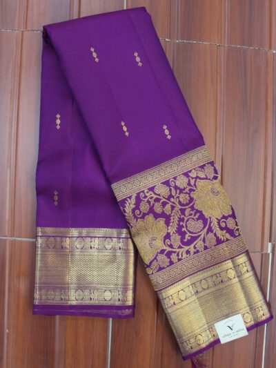 Folded royal purple pure handloom Kanjeevaram silk saree featuring delicate gold buttis, a heavy gold zari border with peacock and geometric mesh patterns, and a grand floral vine zari pallu.