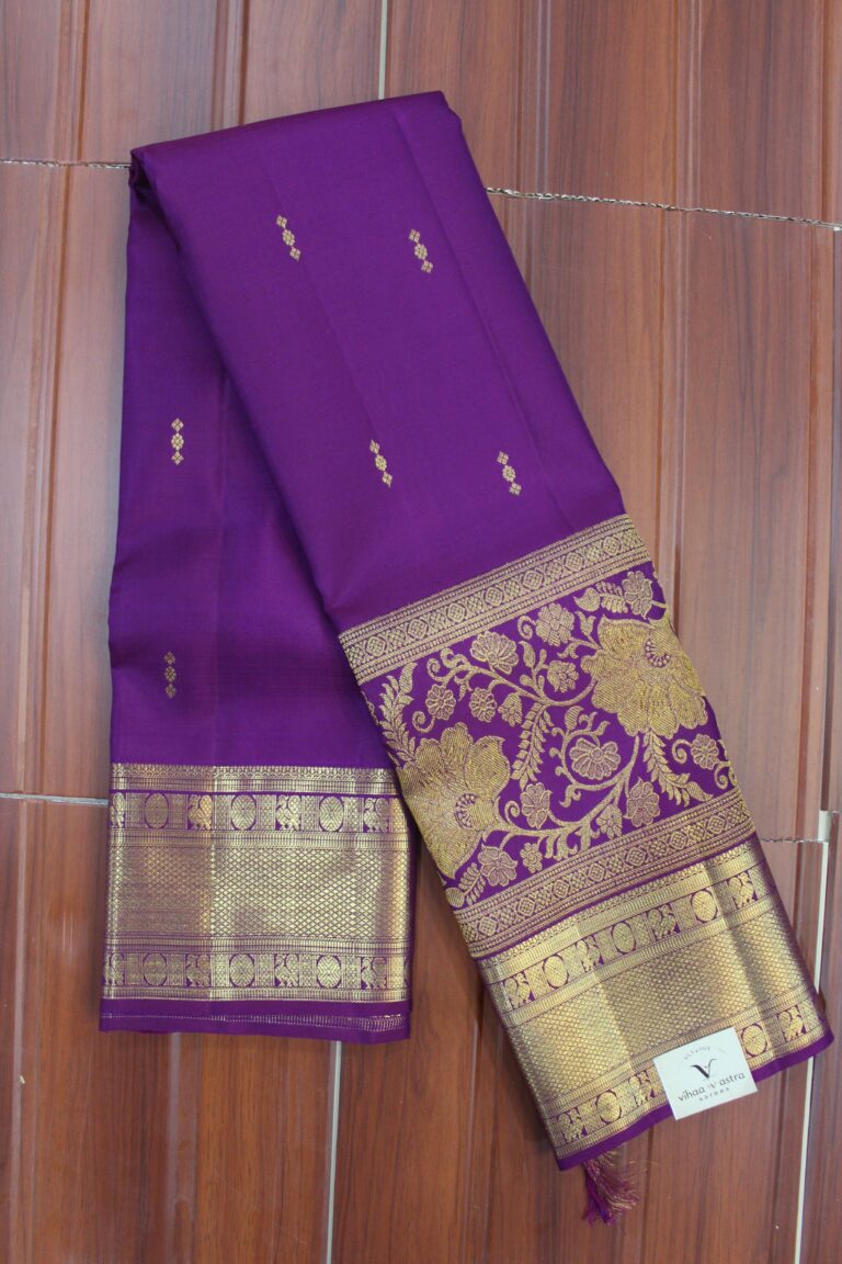 Folded royal purple pure handloom Kanjeevaram silk saree featuring delicate gold buttis, a heavy gold zari border with peacock and geometric mesh patterns, and a grand floral vine zari pallu.