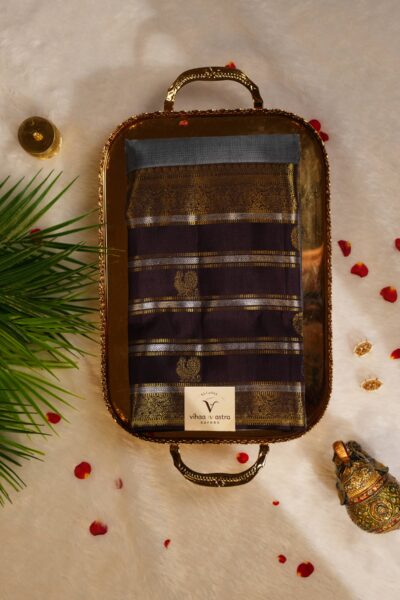Top view of the beautifully folded elephant grey pure handloom Kanjeevaram Pattu saree resting on a decorative brass tray, showcasing the delicate gold stripes and heavy brown border.